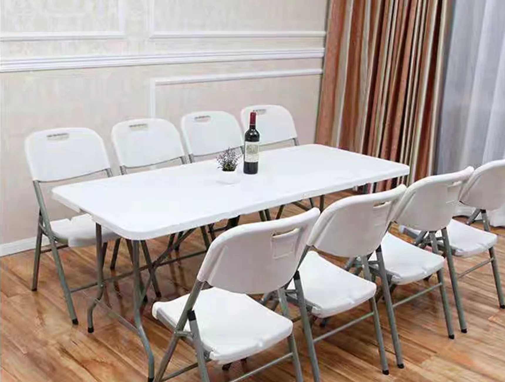 A white folding table with eight white folding chairs arranged around it. A wine bottle and small plant are on the table. The setting is in a room with wooden floors and curtains.