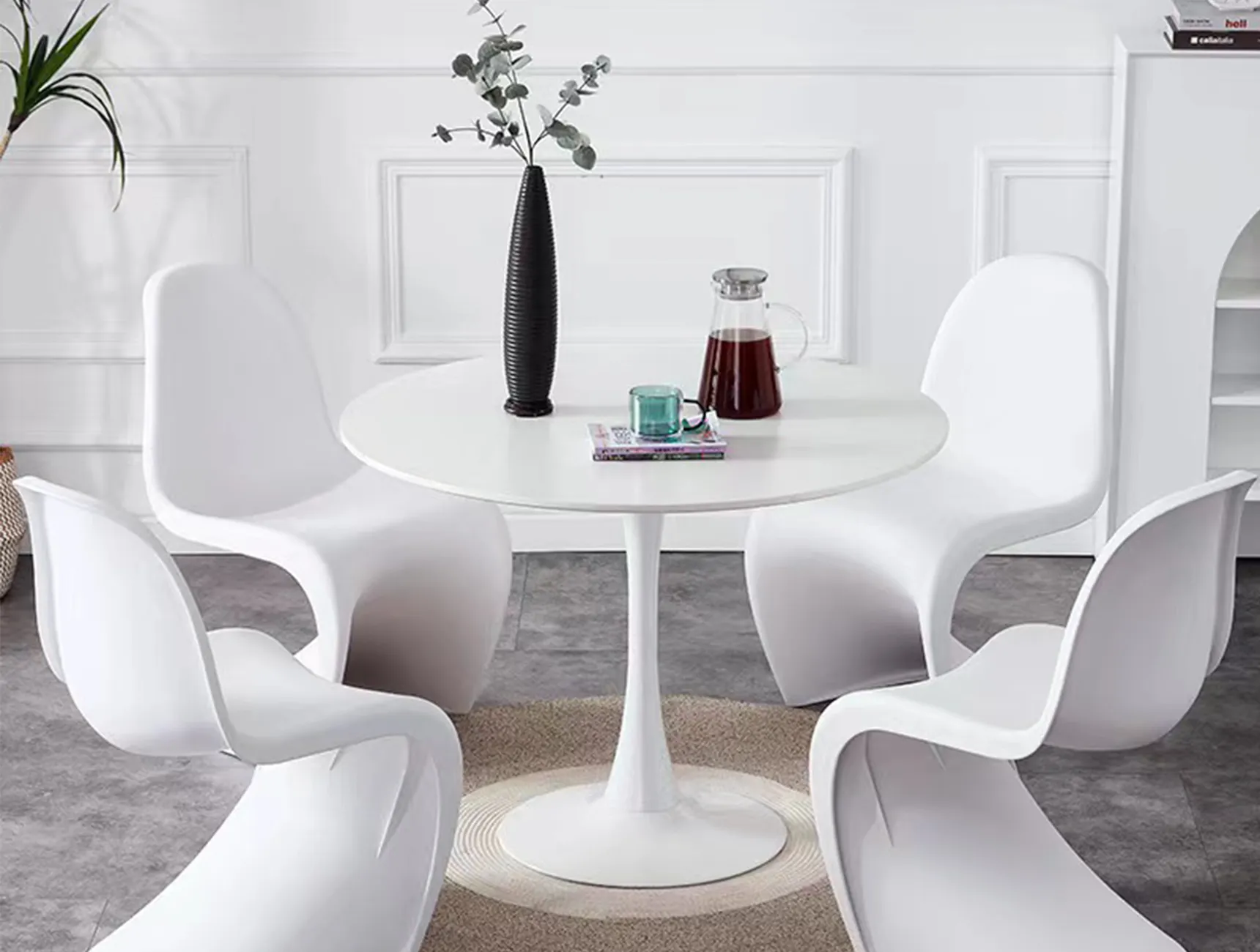 A modern round white table with four white curved chairs. On the table are a tall black vase with plants, a glass carafe with red liquid, and a small teal cup on a book. The setting is in a room with white walls and a gray floor.